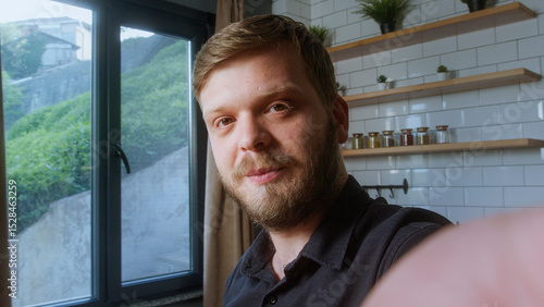 Bearded man looks directly at camera with a gentle smile, suggesting a friendly moment during a video call in natural light.