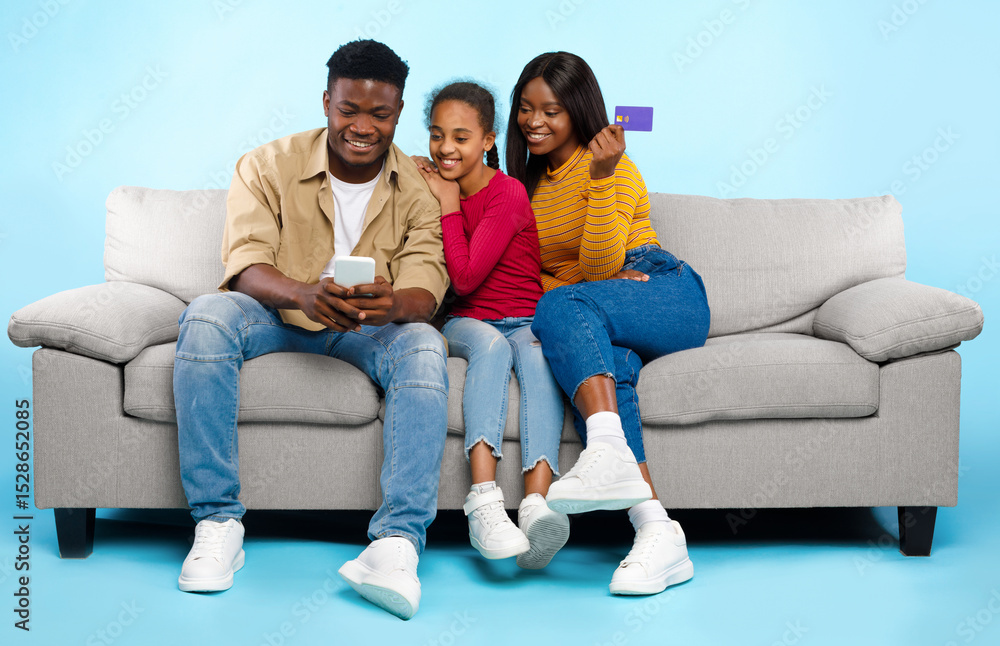 Online Shopping Concept. Happy Young Black Parents With Daughter Using Cell Phone And Debit Credit Card Buying Goods In Internet Web Store, Three People Sitting On Couch Isolated On Blue Studio Wall Online Shopping Concept. Happy Young Black Parents With Daughter Using Cell Phone And Debit Credit Card Buying Goods In Internet Web Store, Three People Sitting On Couch Isolated On Blue Studio Wall
