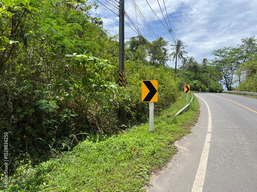 Editorial: Tropical Winding Road Curve Warning Signs. Curved coastal road with yellow and black chevron warning signs indicating sharp turns ahead. Aipril 2025. Krabi,  Thailand 