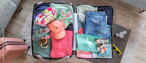 Top view of open suitcase placed on coffee table with colorful summer clothes and accessories, getting ready for vacation, next to vintage photographic camera and yellow sunglasses