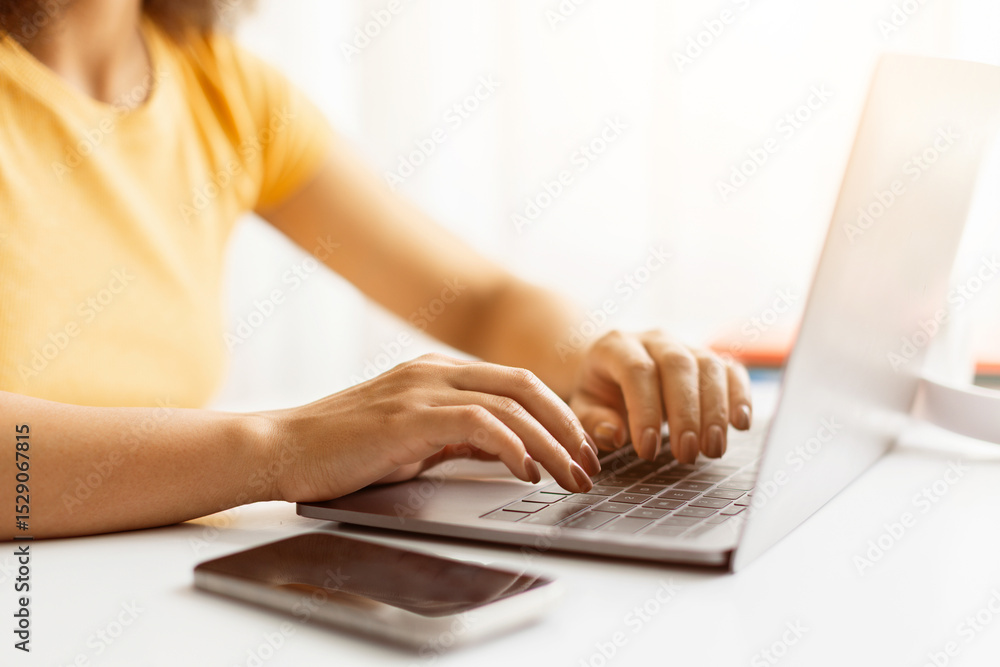 Naklejka premium Hands of young African American woman typing on laptop in office environment, closeup of fingers rest on keyboard, while smartphone lies nearby on the white desk, ideal for studying or remote work