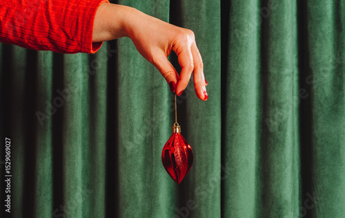 Female hand in red sleeve holds a shiny red Christmas ornament against a green curtain. Minimal and festive holiday concept with vibrant colors and elegance.