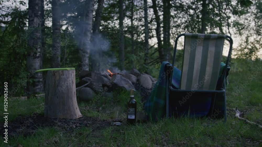 Campfire scene at forest edge with empty striped chair, chopping stump, and bottle on ground at dusk.