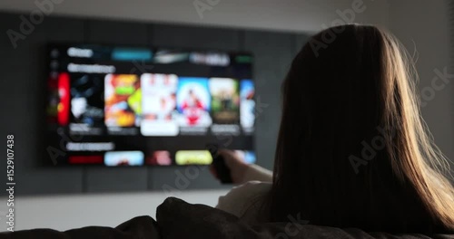Woman selects tv channels with remote control at living room on sofa. Woman controls TV using a modern remote control.