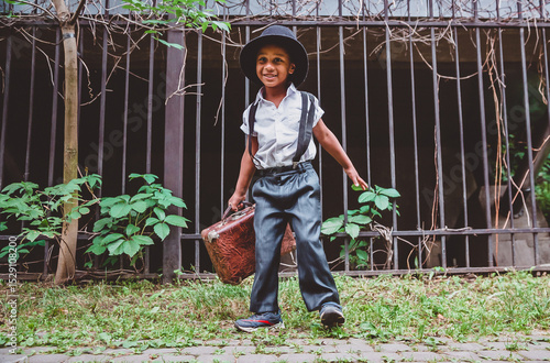 A boy dressed in the style of the 20s with a suitcase in his hand against the background of a fence