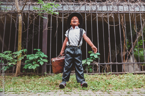 a boy dressed in the style of the 20s with a suitcase in his hand against the background of a fence takes a big breath of fresh air