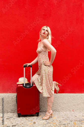 Woman in summer dress travelling with red suitcase in front of a red painted wall