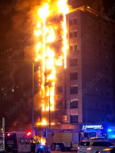 A tall building is engulfed in massive flames, with fire trucks, ambulances, and police vehicles on the street below, responding to the emergency.
