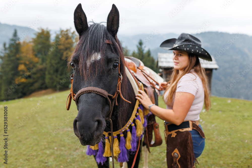 Obraz premium A young woman in western wear stands with her dark horse in a grassy pasture. Mountains and trees are in the background on a cloudy day in Europe.