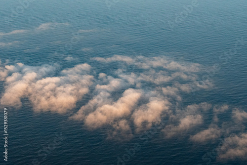 Aerial view showcasing soft, fluffy clouds tinged with warm, golden hues as they hover above the vast, deep blue expanse of the Pacific Ocean. The sunlight creates a gentle contrast