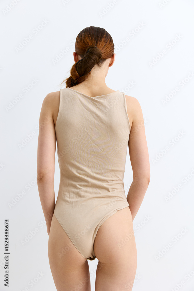 Obraz premium woman in beige bodysuit standing with back turned, red hair tied, minimalistic style, plain white background, natural light, skin exposure