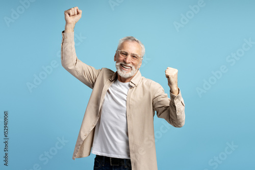 Konstfotografi Euphoric senior man celebrating success with raised fists on blue background