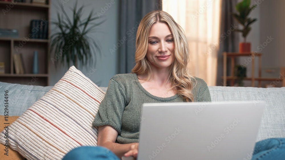 Fototapeta premium Young gorgeous woman typing on laptop sitting on sofa working online. Beautiful busy relaxed female using gadget to communicate. Charming blonde lady in living room. Working from home concept.
