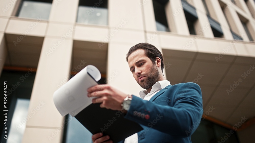 Fototapeta premium Boss of company standing outside near office building. Director holding folder and flipping through business papers. Executive director solving work issues. Leader of company wearing suit and watch.