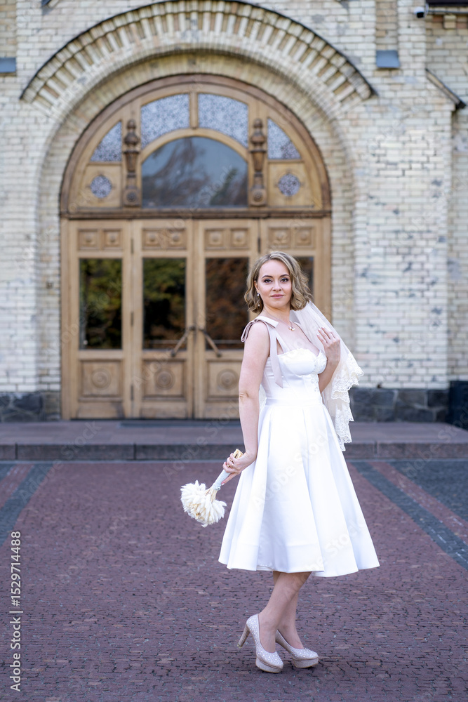 Fototapeta premium full length portrait of beautiful young bride in white short wedding dress and veil. Bride holding bouquet of dried flowers. Wedding day