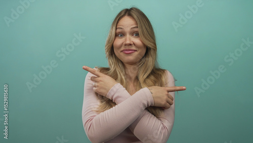 Blonde woman pointing with crossed wrists and index fingers in teal studio setting; indecision confusion.