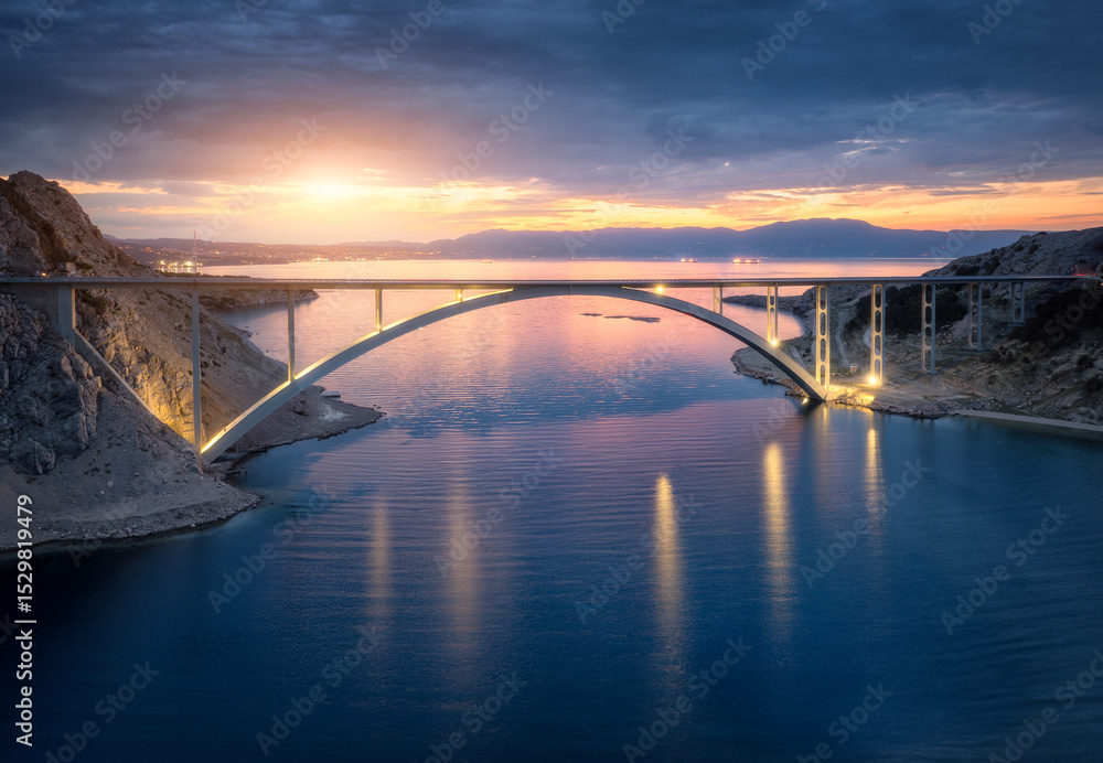 Fototapeta premium Aerial view of bridge and blue sea at night. Modern Krk bridge with illumination, island, mountains, water and cloudy sky at sunset. Top drone view of road, city lights, rocks at dusk. Architecture