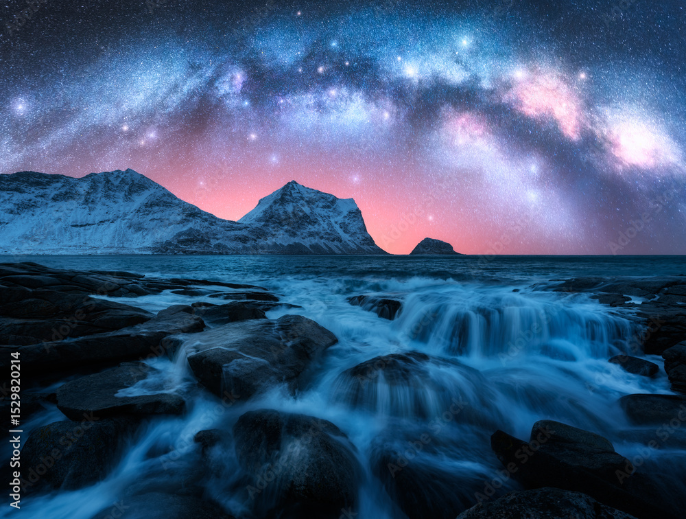 Naklejka premium Night starry sky with glowing Milky Way, rocky shore with stones in water. Winter colorful landscape with milky way and stars, fjord, pink sky, sea, snowy mountains in Lofoten Islands, Norway. Space