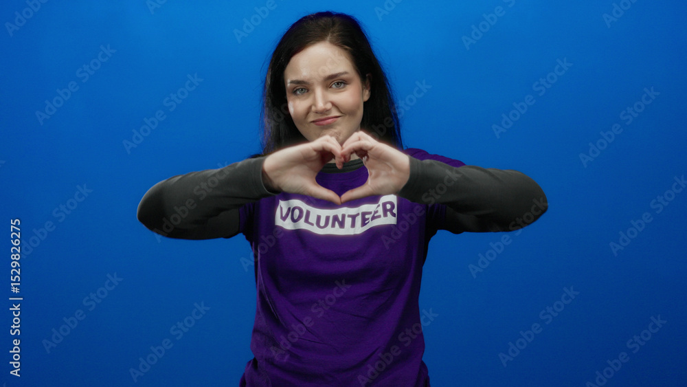 Naklejka premium Brunette woman in purple volunteer shirt gestures heart with hands against vibrant blue background, conveying emotion and dedication.