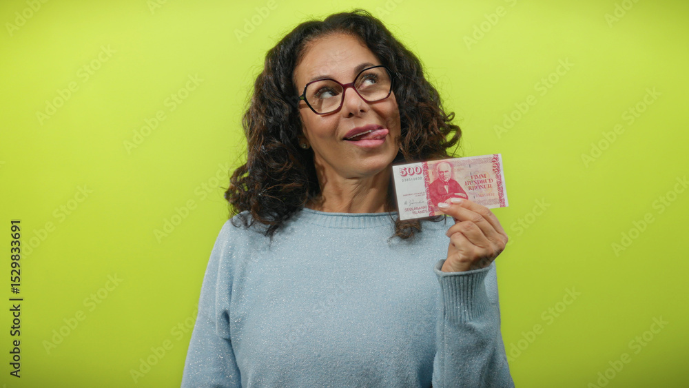 Middle-aged woman shows icelandic krona banknote with playful expression against a vibrant yellow background, highlighting the concept of finance and currency with a touch of humor. Middle-aged woman shows icelandic krona banknote with playful expression against a vibrant yellow background, highlighting the concept of finance and currency with a touch of humor.