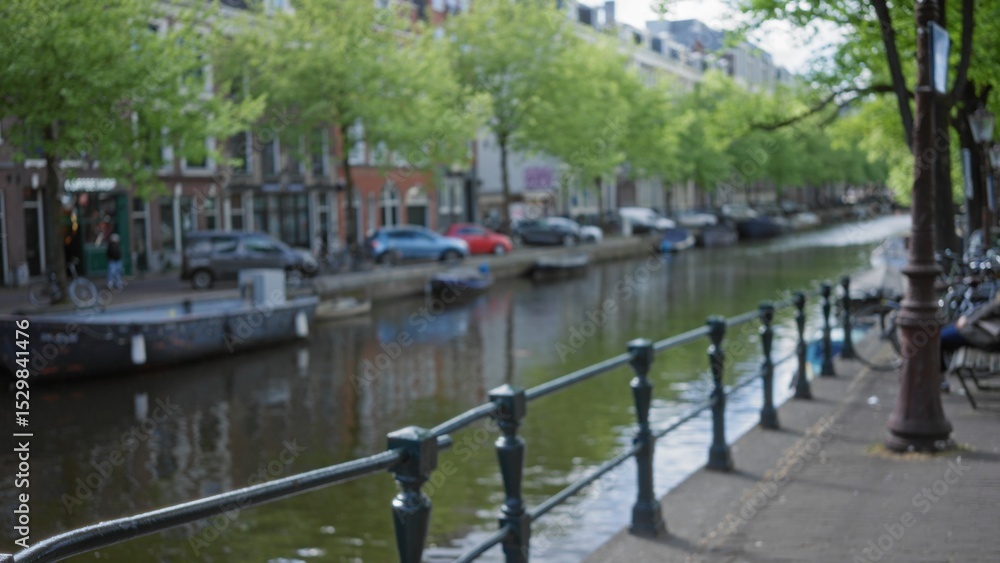 Fototapeta premium Blurred view of amsterdam canals with trees, buildings, and boats reflecting in the water creating a serene, peaceful atmosphere with a defocused, bokeh effect.