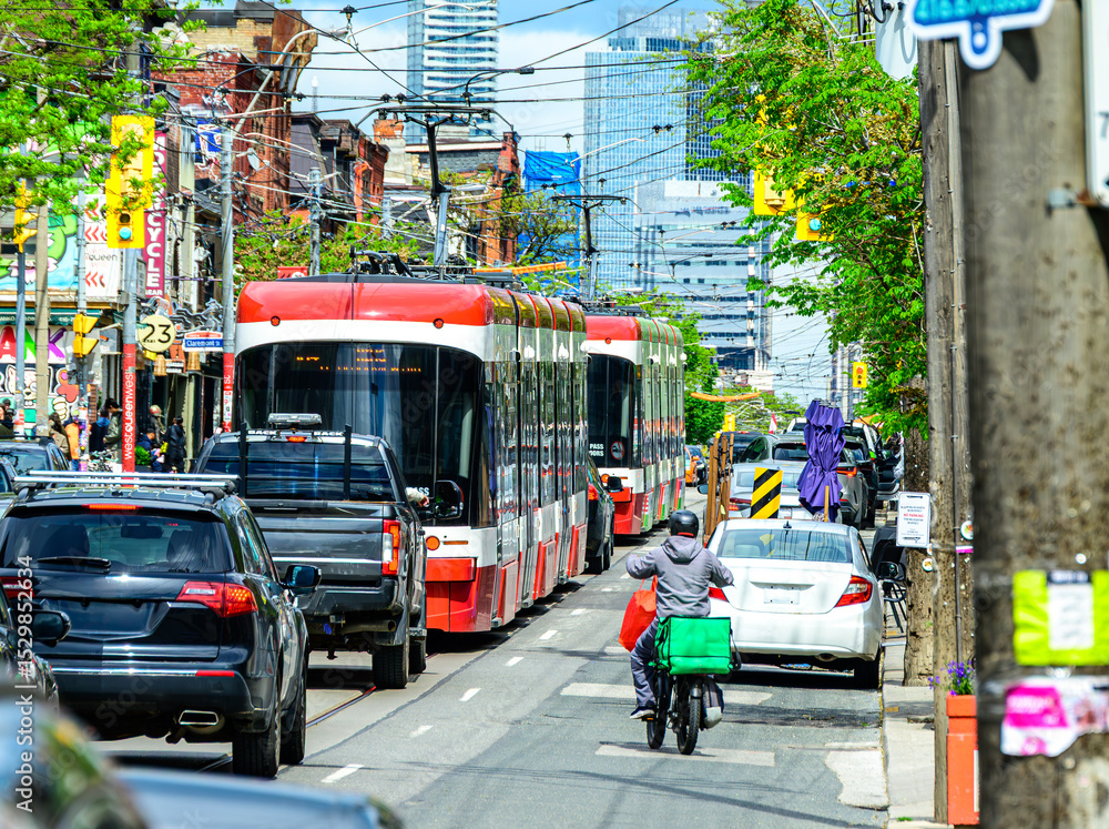 Fototapeta premium Gig economy: food delivery driver on aan electric bike in busy traffic with cars and light rail street cars shot on a sunny day in Toronto's Queen West district in summer