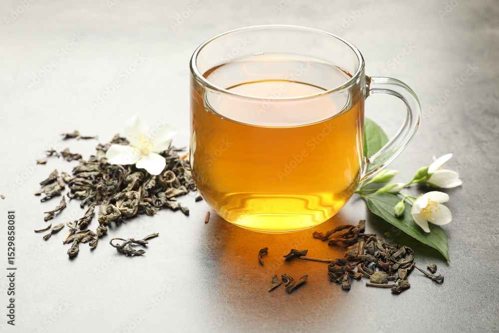 Fototapeta premium Aromatic jasmine tea in glass cup, brew and flowers on grey table, closeup