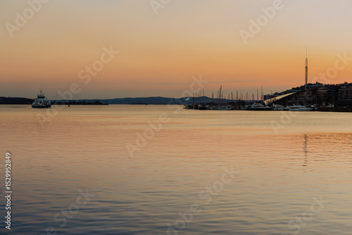 Photography Bathed in the warm glow of sunset, Oslo harbour showcases traditional colourful wooden houses along the waterfront, with boats gently berthed in the distance