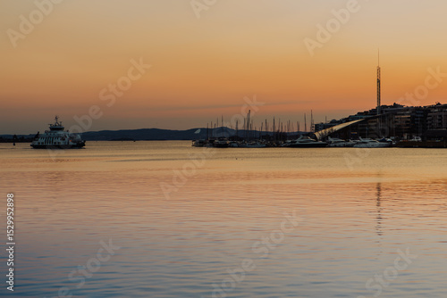 Photography Bathed in the warm glow of sunset, Oslo harbour showcases traditional colourful wooden houses along the waterfront, with boats gently berthed in the distance