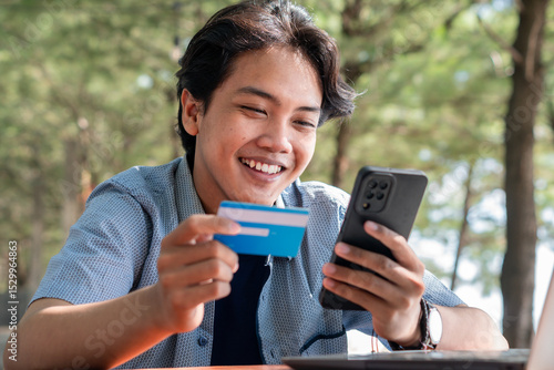 Happy Asian Young businessman hold blue creditcard and phone purchasing online shopping in outdoor cafe.