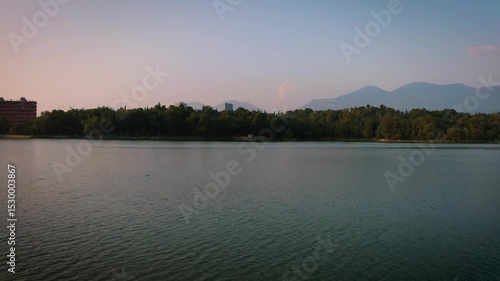 Forward drone footage starting over Tirana’s artificial lake at sunset, rising to reveal the city skyline framed by trees and distant mountains in the capital of Albania.