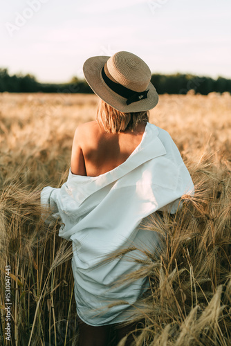 Back view woman in straw boater and white off-shoulder shirt standing in golden wheat field at sunset