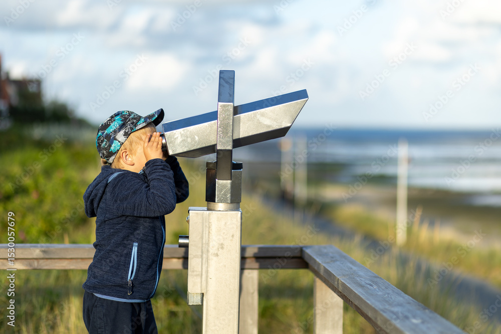 Fototapeta premium Child observes scenic landscape through binoculars on a beach boardwalk during the day