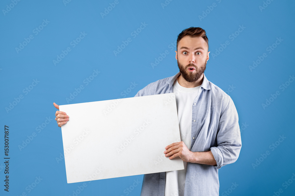 Surprised young man showing blank white poster, presenting something on blue studio background, space for design. Hipster guy demonstrating empty banner, mockup for your advertisement Surprised young man showing blank white poster, presenting something on blue studio background, space for design. Hipster guy demonstrating empty banner, mockup for your advertisement
