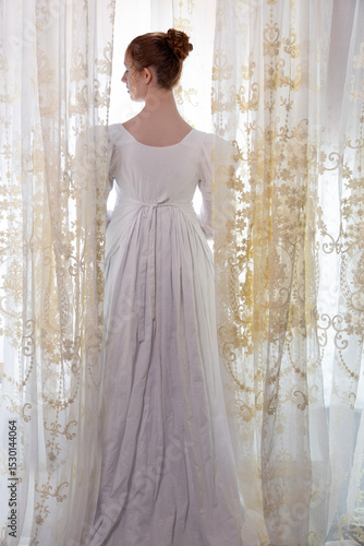 A Regency period woman wearing a white embroidered cotton dress and surrounded by backlit lace curtains