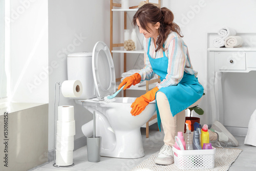 Young woman cleaning toilet bowl with brush in light bathroom