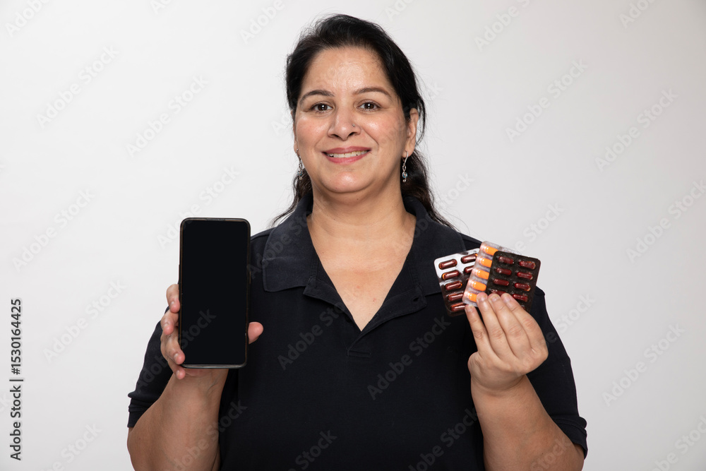 Indian middle age smiling woman holding smartphone with medicine pills over isolated grey studio background Indian middle age smiling woman holding smartphone with medicine pills over isolated grey studio background