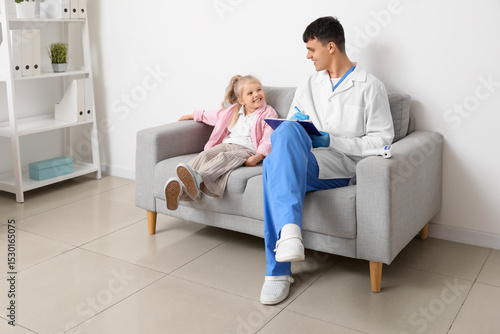 Male pediatrician with clipboard and little girl sitting on sofa in clinic