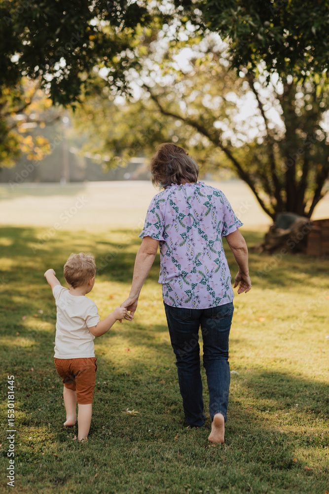 Fototapeta premium Grandmother Walking Hand in Hand with Young Grandson in Sunny Park, Outdoor Family Bonding Moment Captured from Behind