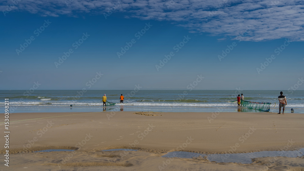 Obraz premium A group of people is pulling fishing nets out of the ocean. Turquoise waves spread across the sand. Reflection. Low tide. Puddles of water in the foreground. Blue sky, clouds. Copy space. Madagascar. 
