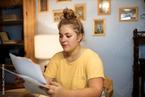 Indoor image of concerned woman in casual yellow t-shirt with hair knob examining paper documents sitting at home in living-room, reading rent agreement or loan statement from bank