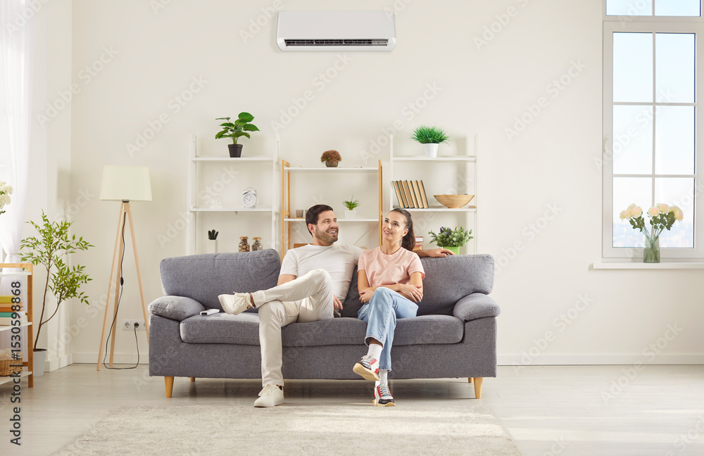 Obraz premium Happy couple sitting on comfortable gray sofa in living room, relaxing together under air conditioner. Smiling young man and woman resting on couch, talking and enjoying fresh cool air at home.