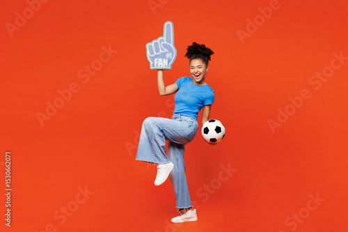 Fototapeta Naklejka Na Ścianę i Meble -  Full body young woman fan she wear blue t-shirt foam 1 glove finger up cheer up support football sport team hold soccer ball watch tv live stream raise up leg isolated on plain red orange background.