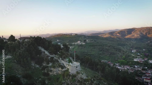 Pullback drone footage from Berat Castle’s balcony at golden hour, slowly revealing the surrounding hills, scattered homes, and lush countryside of central Albania.