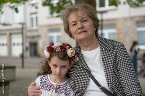Sad granddaughter in a traditional Ukrainian embroidered shirt and flower wreath sitting beside her loving grandmother on a bench in summer. Concept of childhood and generational bond