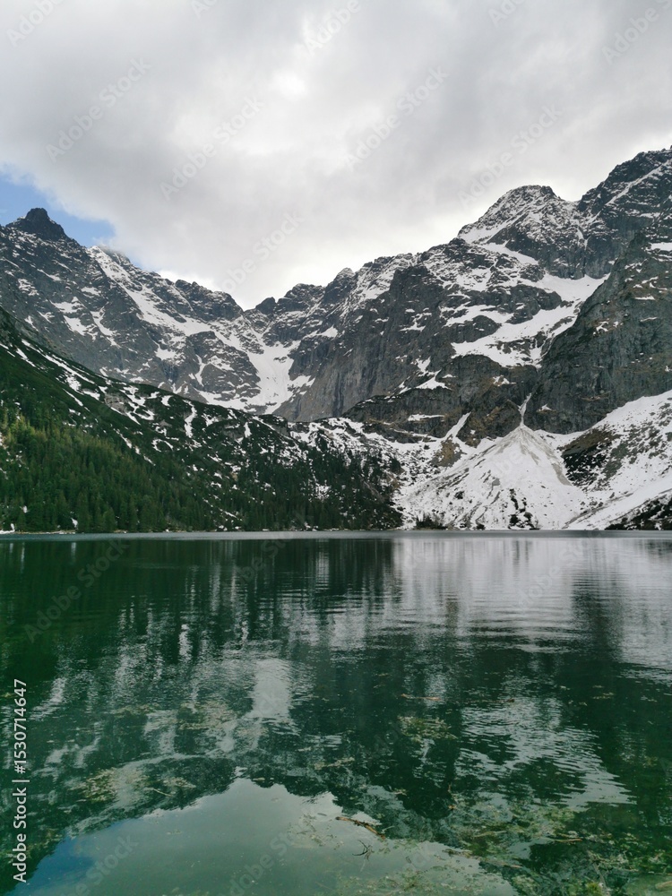 Towering snow-covered Tatra Mountains are reflected in the clear emerald waters of Morskie Oko lake beneath a moody sky near Zakopane, Poland Towering snow-covered Tatra Mountains are reflected in the clear emerald waters of Morskie Oko lake beneath a moody sky near Zakopane, Poland