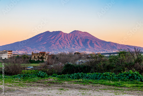 A breathtaking view of Mount Vesuvius during sunset, with soft orange and pink hues illuminating the majestic volcano. The foreground features rural ruins and greenery, creating a serene and timeless 