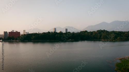 Rising drone footage over Tirana’s Grand Park lake, gradually revealing the city skyline and its modern skyscrapers in the background, set against a calm sunset atmosphere in Albania.
