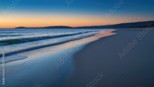 Fototapeta Naklejka Na Ścianę i Meble -  A tranquil beach at sunset, where golden hues merge into a wide blue sky. Soft waves ripple onto the shore, and footprints in the sand trail toward the horizon, evoking a peaceful, everlasting beauty.