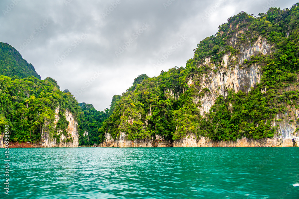 View of Khao Sok Lake in Krabi province, Thailand, with towering limestone cliffs covered in tropical rainforest rising above the calm surface of the lake. View of Khao Sok Lake in Krabi province, Thailand, with towering limestone cliffs covered in tropical rainforest rising above the calm surface of the lake.